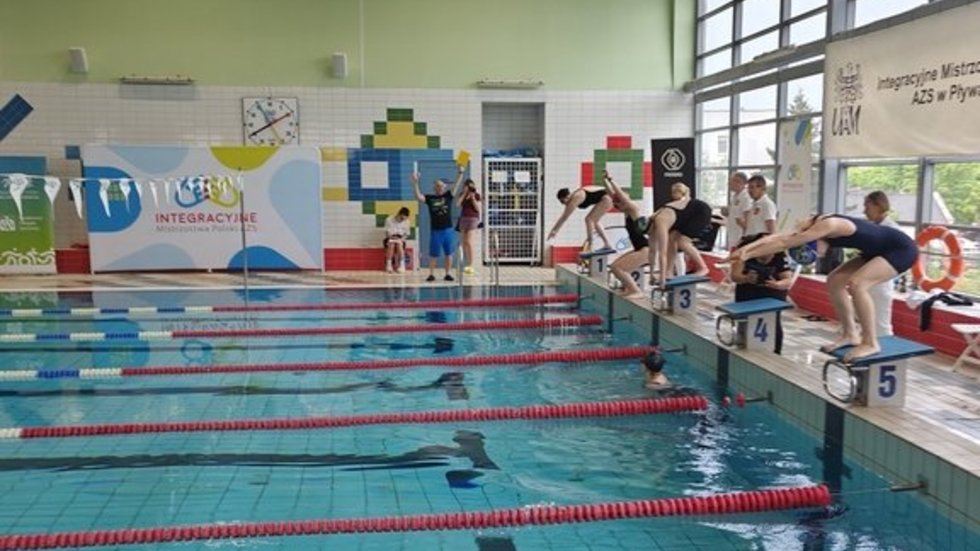 Wnętrze krytej pływalni podczas zawodów pływackich. Kilku zawodników w strojach kąpielowych stoi na słupkach startowych, przygotowując się do skoku do wody. Jeden z zawodników jest już w trakcie skoku.